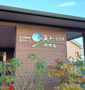 木調の外壁に施設名看板がある建物外観。玄関周辺の景観。